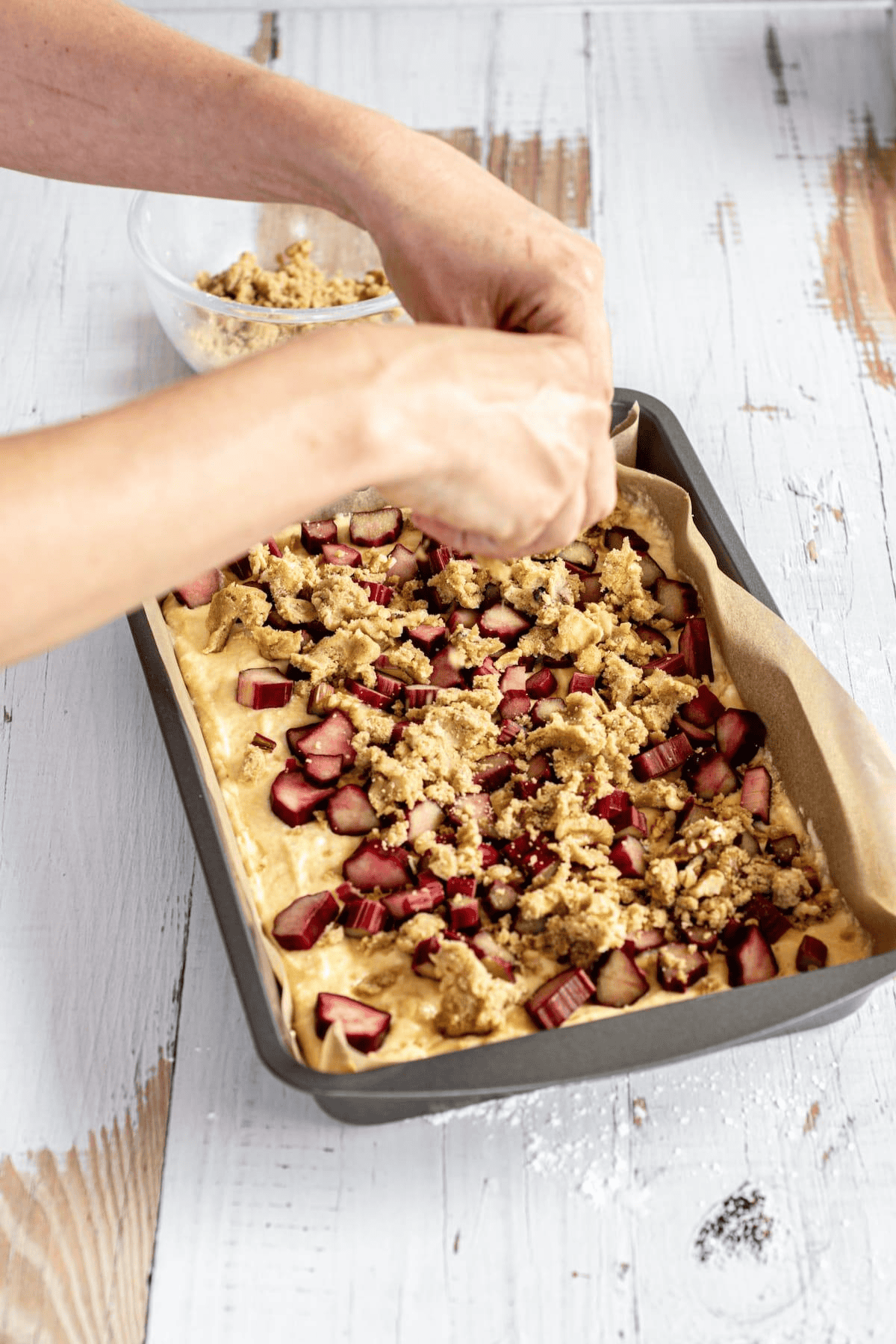 A person sprinkles crumb topping over a rectangular baking tray filled with chopped rhubarb and batter, preparing a rhubarb streusel cake on a white wooden surface. A bowl of extra crumb topping sits nearby.