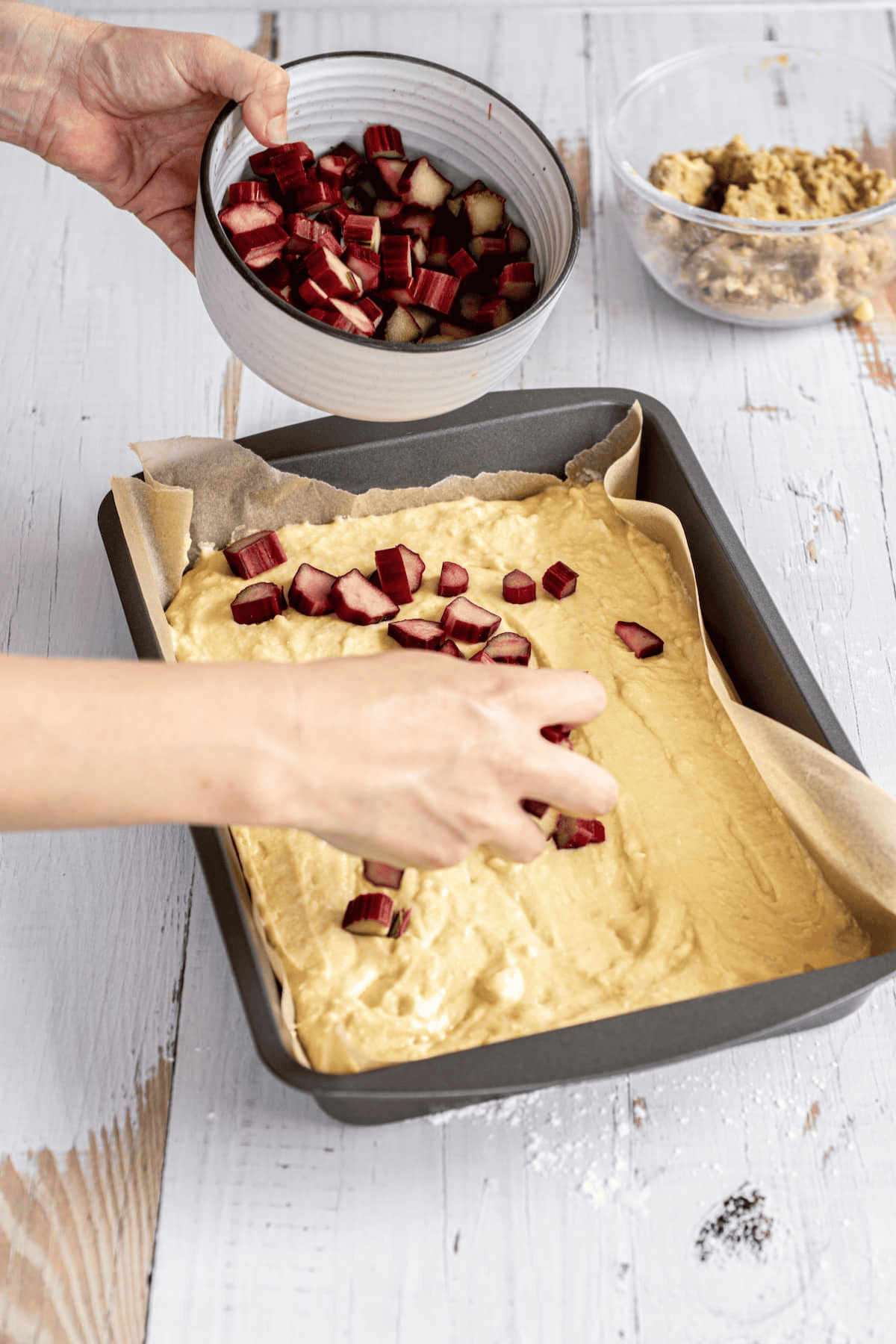 A person sprinkles chopped rhubarb onto cake batter in a rectangular baking pan lined with parchment, preparing a delicious rhubarb streusel cake; nearby are bowls of chopped rhubarb and dough on a white wooden table.