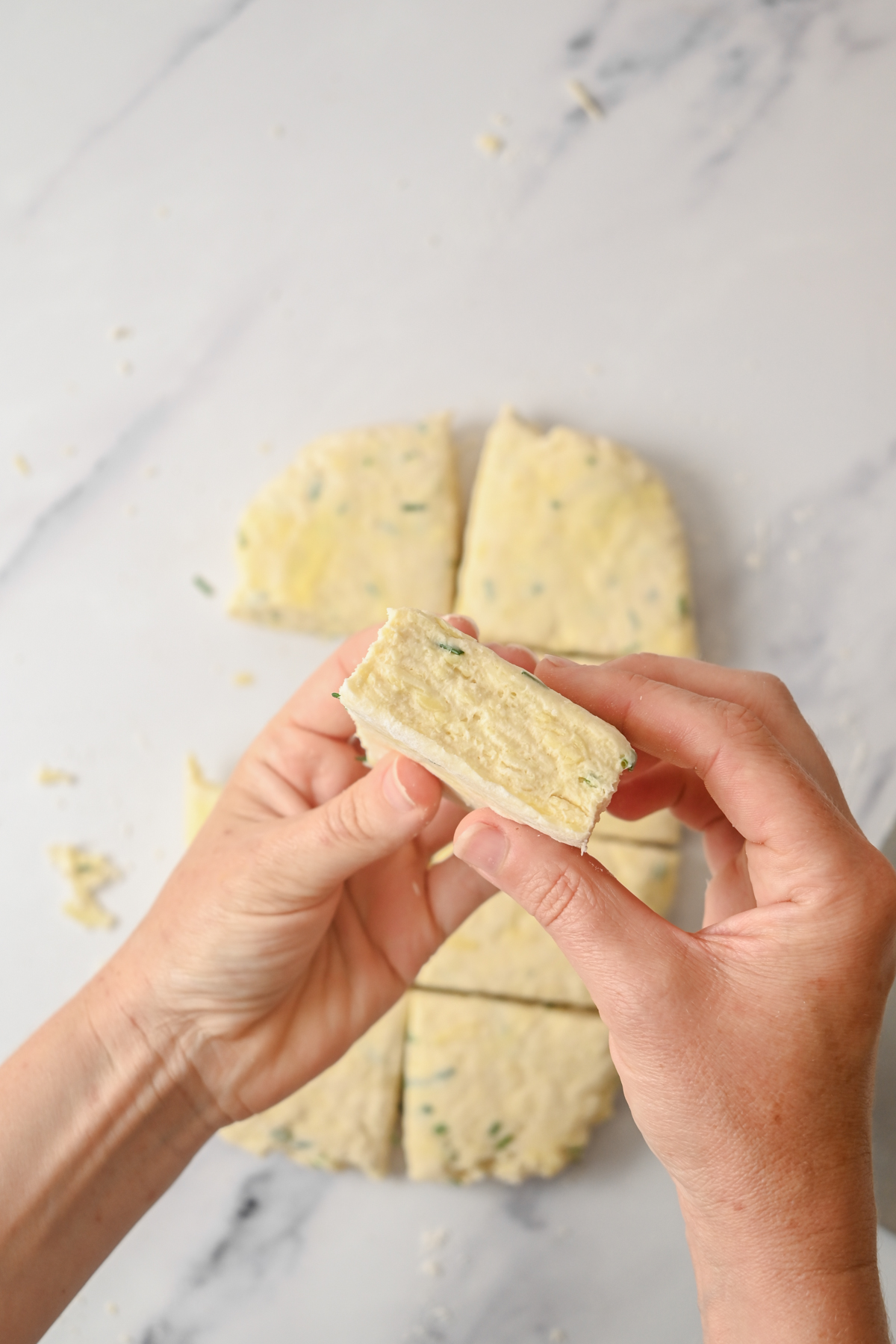 Two hands hold a triangular piece of unbaked sourdough cheese scone dough, showing its fluffy texture. More dough pieces are visible on a marble surface in the background.