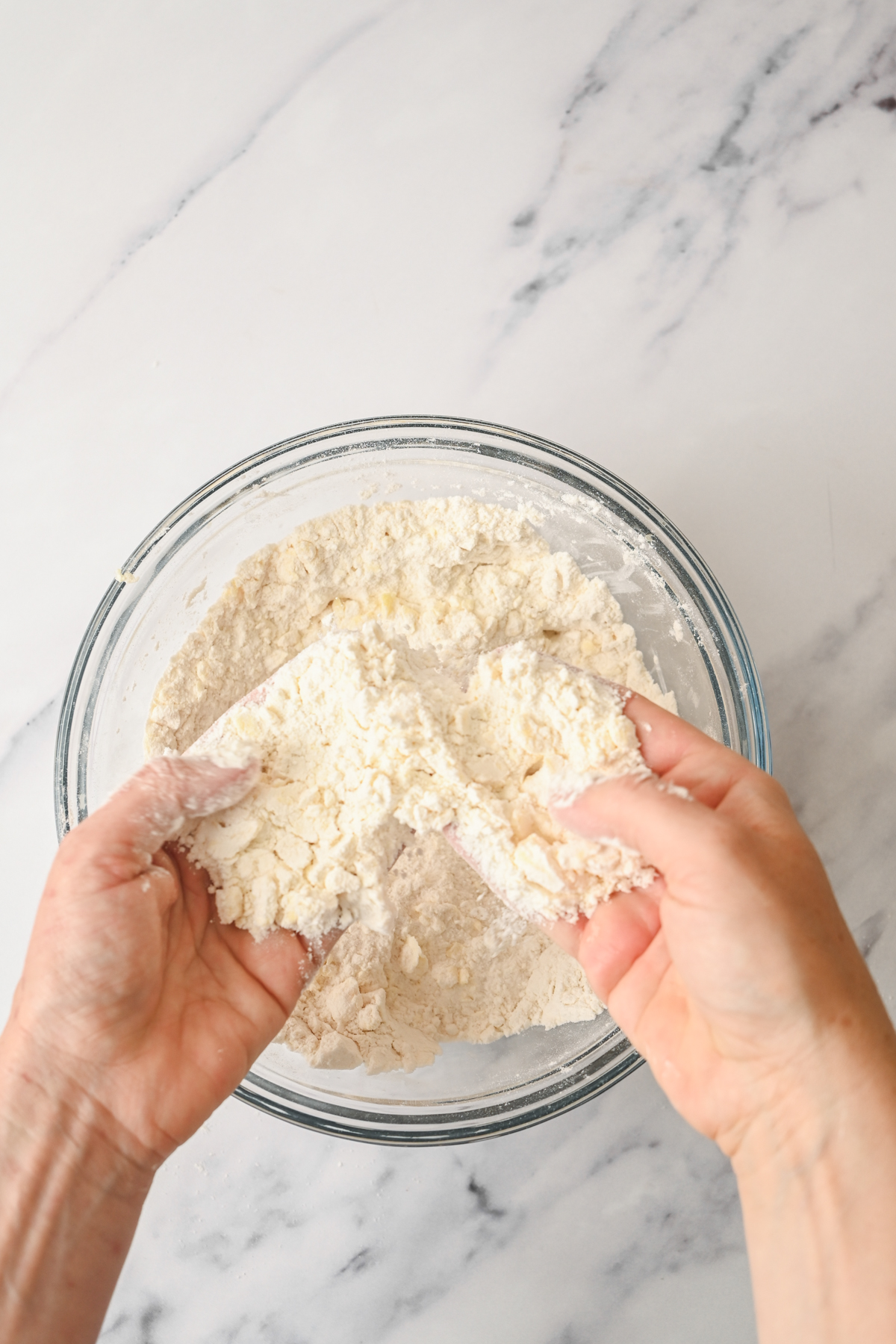 Two hands are mixing flour and butter in a glass bowl on a marble countertop, rubbing them together to create coarse crumbs for sourdough cheese scones.