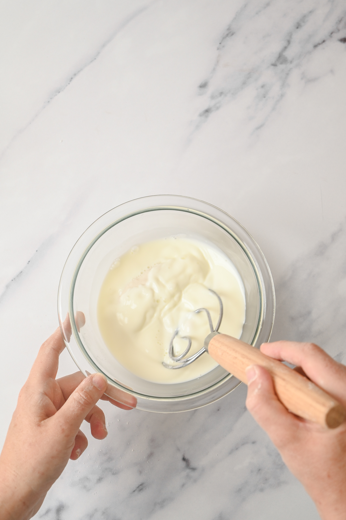 A person uses a metal whisk with a wooden handle to mix thick, white yogurt or cream in a clear glass bowl on a white marble countertop, preparing the base for delicious sourdough cheese scones.