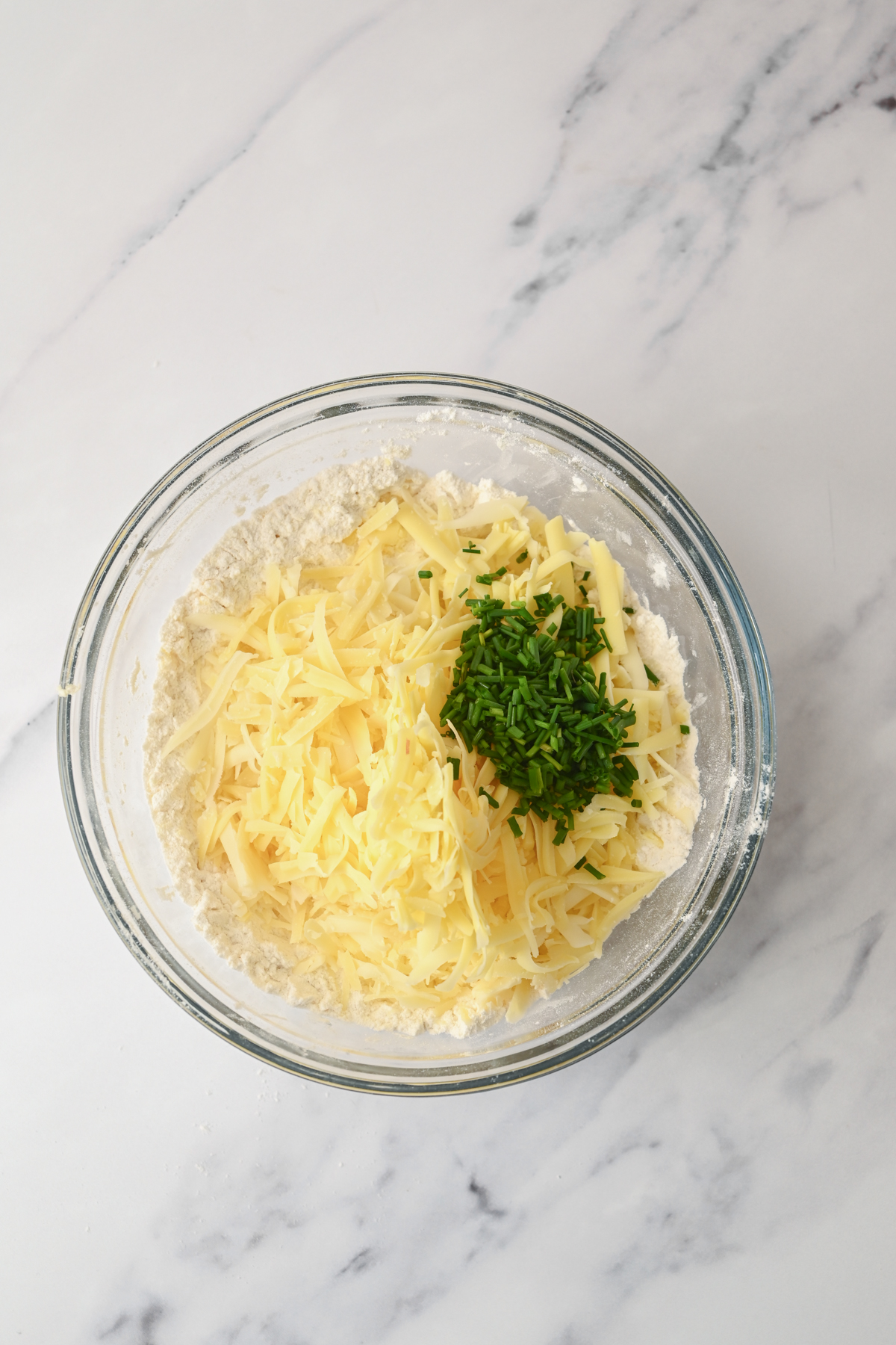 A glass bowl on a marble surface contains grated cheese and chopped chives on top of a white flour mixture, ready to be mixed together for delicious sourdough cheese scones.