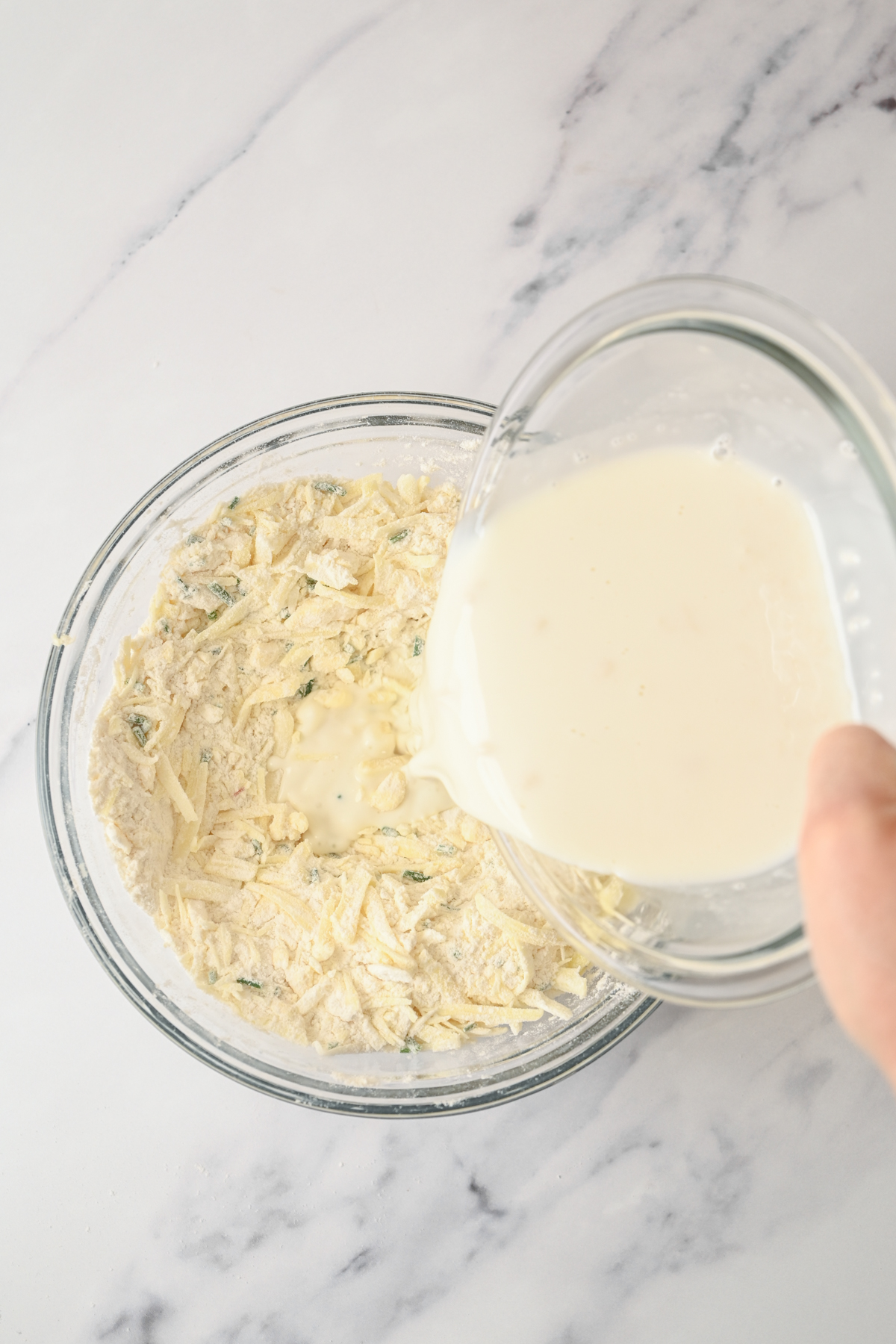 A hand pours milk from a glass bowl into a larger bowl of flour, shredded cheese, and herbs—ingredients for delicious sourdough cheese scones—all set on a white marble surface.