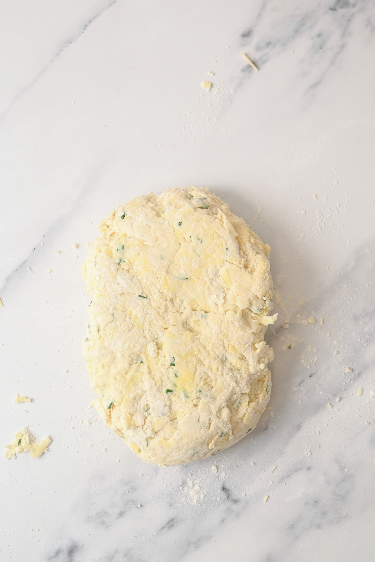 A rough, oval-shaped ball of dough for sourdough cheese scones, speckled with green herbs, sits on a floured white marble surface.