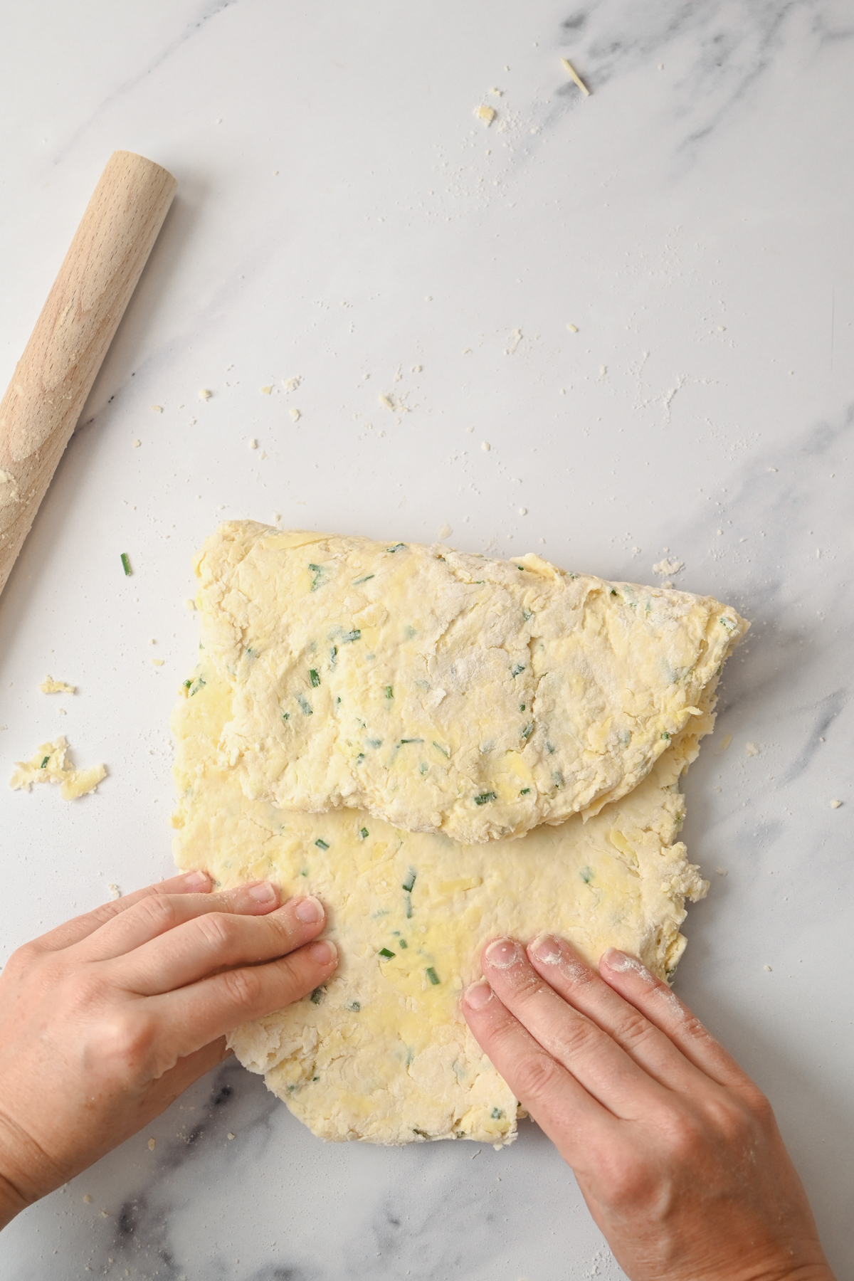 Hands fold a rectangular sheet of dough for sourdough cheese scones with green herbs on a lightly floured marble surface, next to a wooden rolling pin.