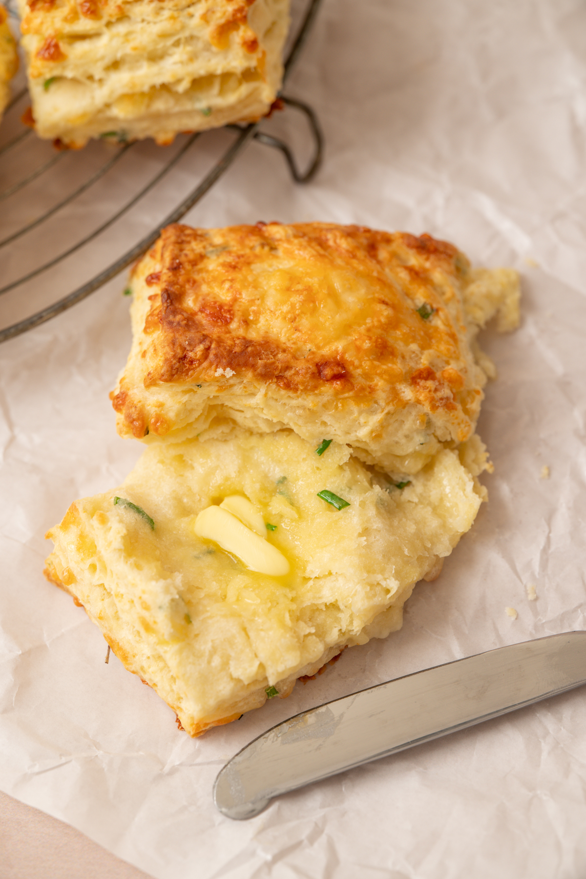 A flaky cheddar and chive biscuit, inspired by classic sourdough cheese scones, is split open with a pat of melting butter on top. It sits on parchment paper next to a metal butter knife, with more biscuits cooling in the background.