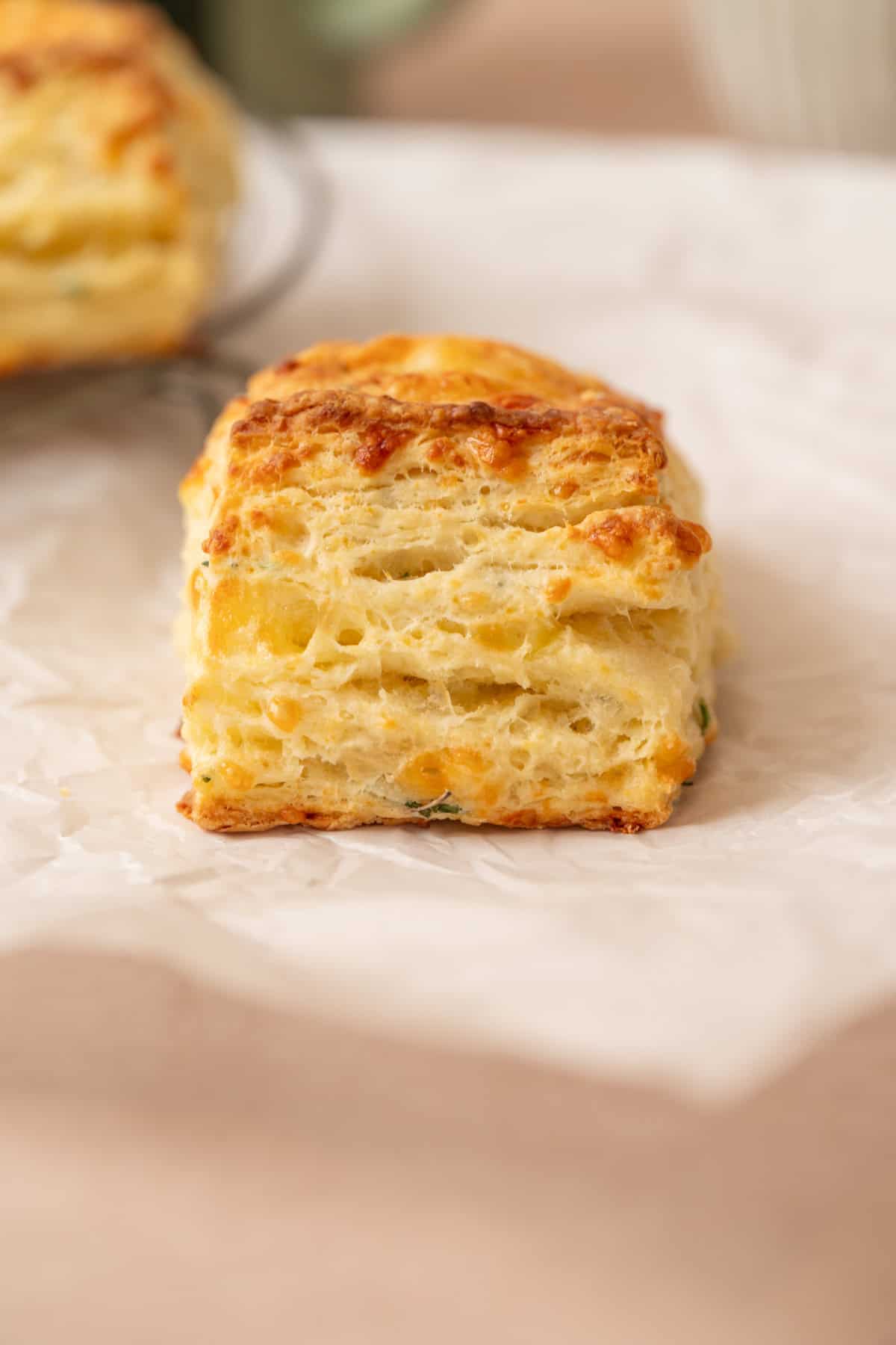 A close-up of golden, flaky sourdough cheese scones with visible layers, resting on a sheet of white parchment paper.