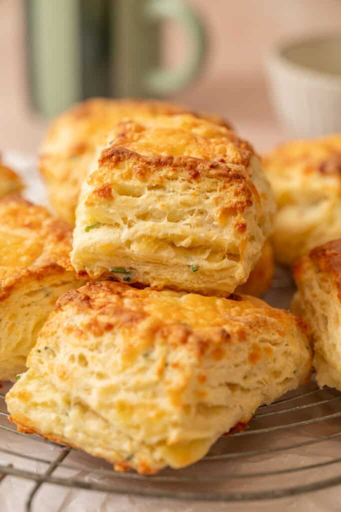 A close-up of golden, fluffy, square buttermilk biscuits and sourdough cheese scones stacked on a cooling rack, showing their layered, flaky texture.