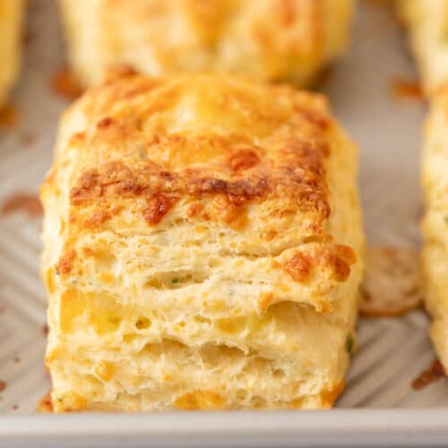 A close-up of a golden, flaky sourdough cheese scone with visible layers, resting on a parchment-lined baking sheet. The scone appears freshly baked with a slightly crisp, cheesy top.