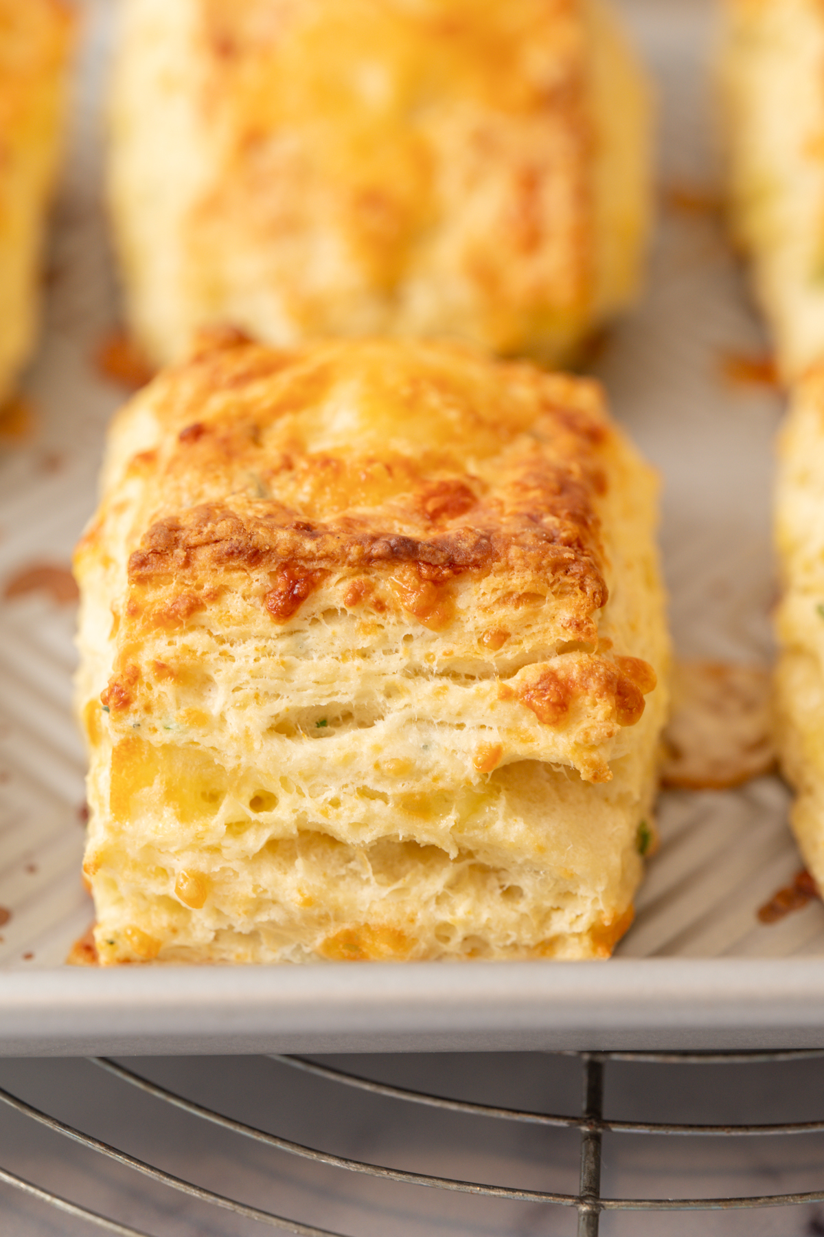 A close-up of a golden, flaky sourdough cheese scone with visible layers, resting on a parchment-lined baking sheet. The scone appears freshly baked with a slightly crisp, cheesy top.