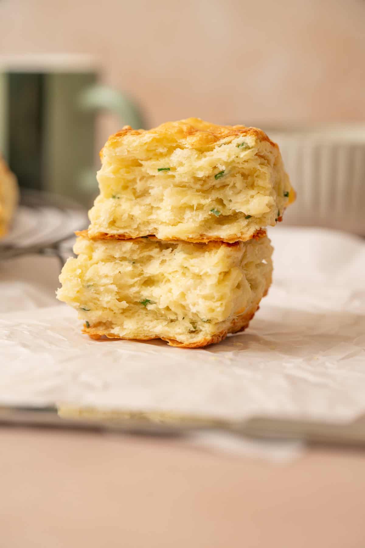 Two flaky, golden-brown sourdough cheese scones with herbs are stacked on white parchment paper. The scones have a soft, layered texture, and a blurred mug and plate are in the background.