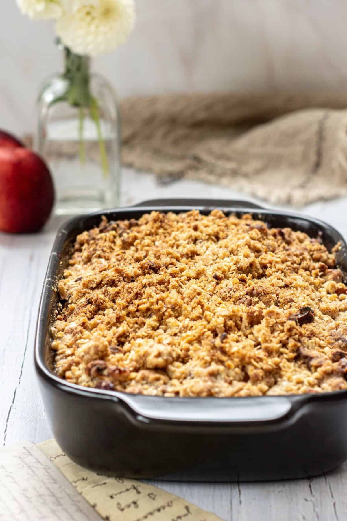 A black baking dish filled with golden-brown apple crisp sits on a light wooden table, reminiscent of a Peach Crisp Recipe With Oatmeal. Apples, a vase of white flowers, and a burlap cloth complete the cozy backdrop.