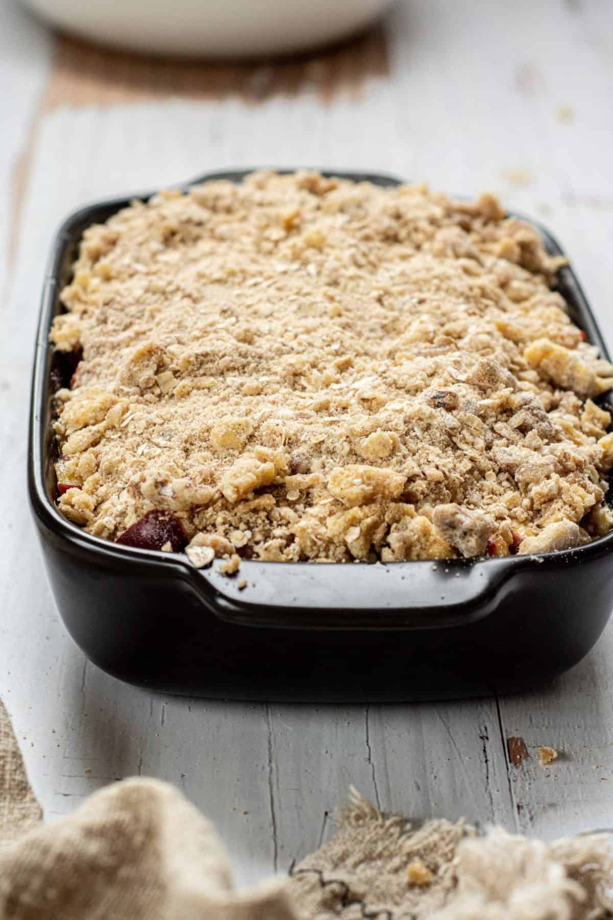 A rectangular black baking dish filled with an unbaked Peach Crisp Recipe With Oatmeal, topped with a thick layer of crumbly, golden streusel, sits on a rustic white wooden surface.