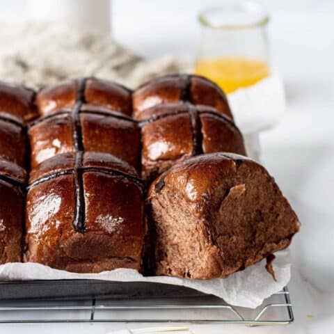 A batch of shiny Orange Chocolate Hot Cross Buns, with one bun pulled aside to reveal a soft, fluffy interior. The buns rest on parchment paper atop a cooling rack, with a glass of orange juice in the background.