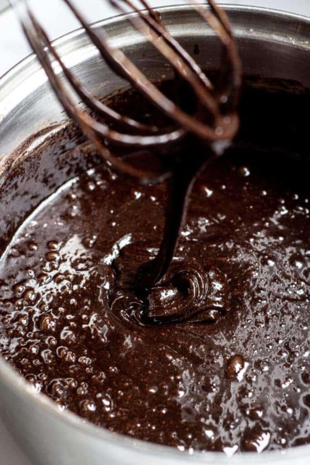 A close-up of a metal mixing bowl filled with glossy triple chocolate brownie batter being mixed, with a whisk lifted above the batter and drips falling back into the bowl.