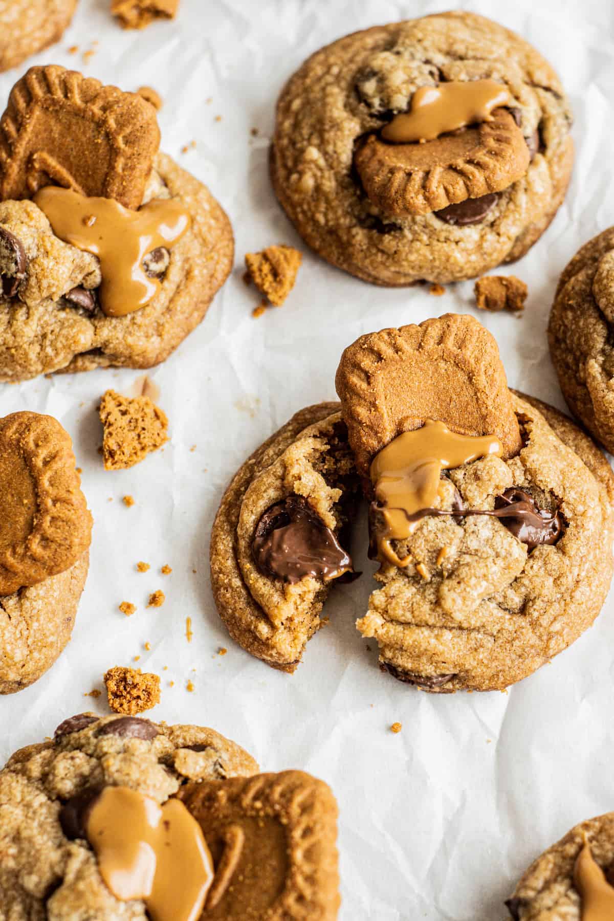 Chocolate chip cookies topped with caramel-drizzled biscoff butter cookies, some whole and some broken to reveal melty chocolate inside, are scattered on a sheet of parchment paper with cookie crumbs around.