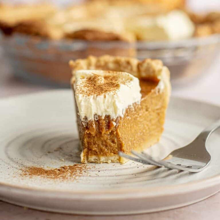 A slice of pumpkin pie without evaporated milk, topped with whipped cream and sprinkled with cinnamon, sits on a white plate, with a fork taking a bite. The rest of the pie is visible in the background.