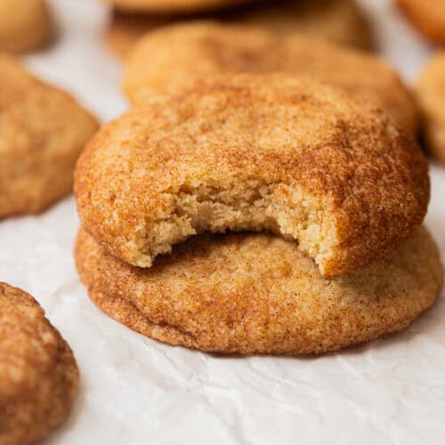 A close-up of a stack of Small-Batch Snickerdoodles on crinkled parchment paper, with the top cookie missing a bite. Other cookies are visible in the background.