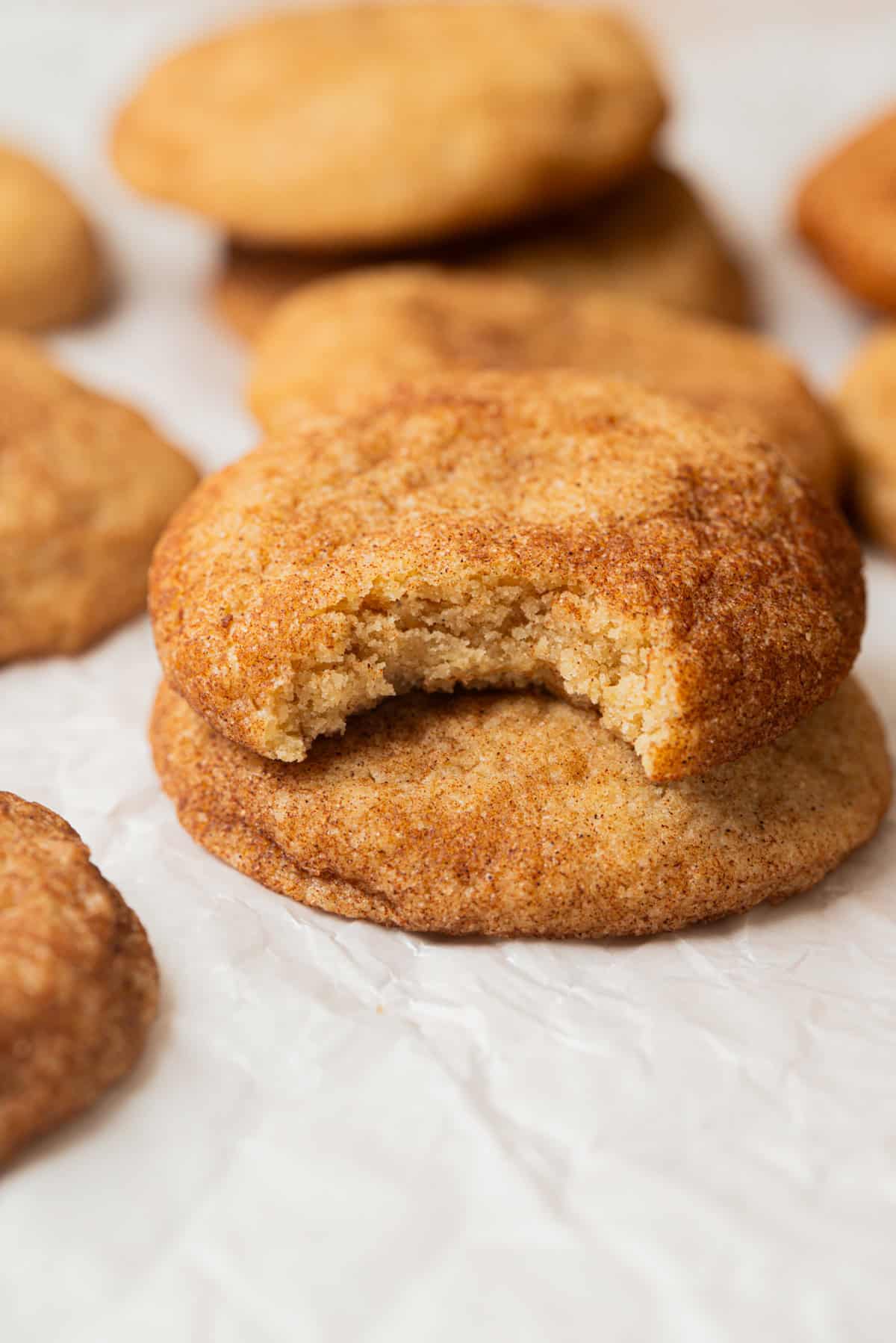 A close-up of a stack of Small-Batch Snickerdoodles on crinkled parchment paper, with the top cookie missing a bite. Other cookies are visible in the background.