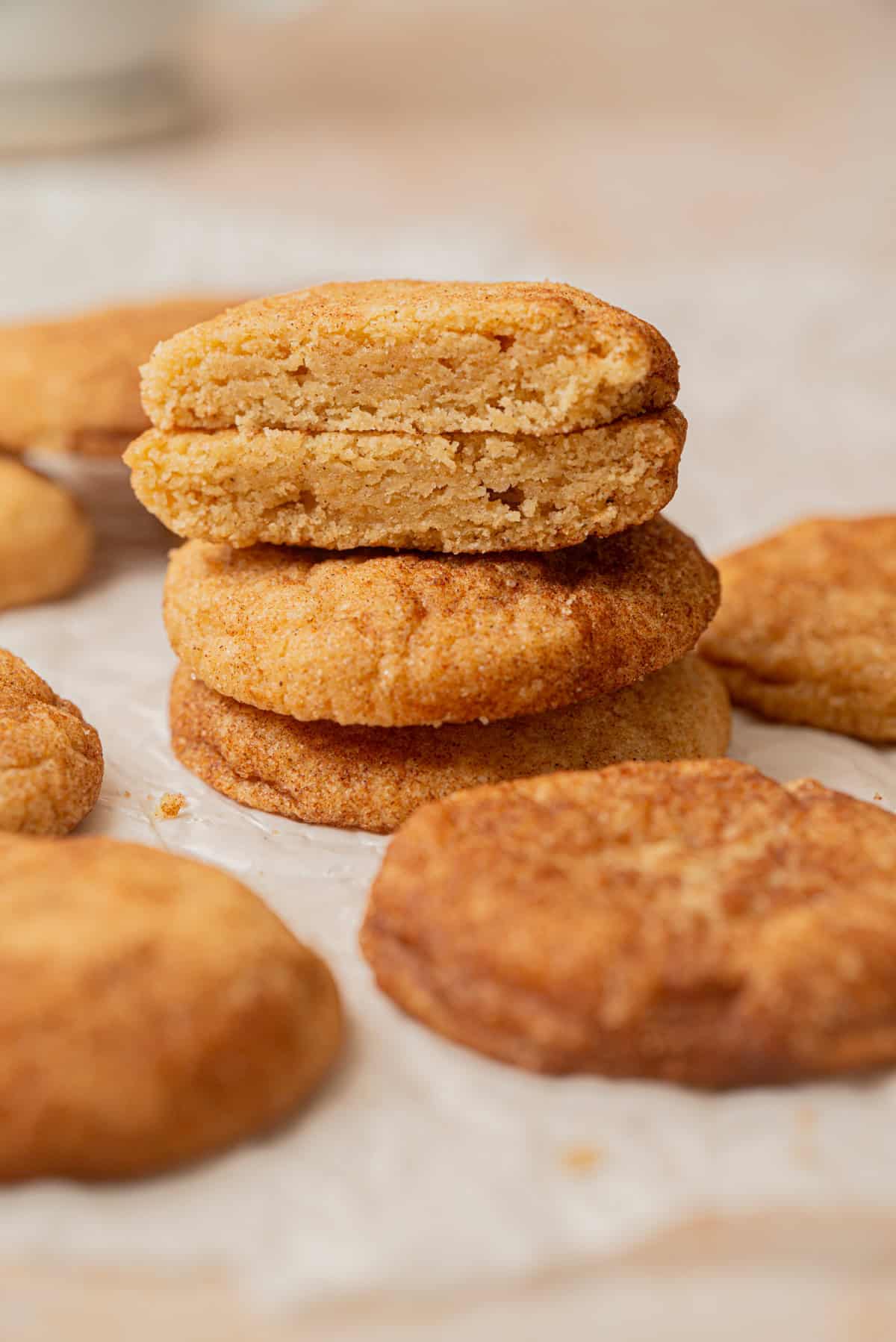 A close-up of golden brown Small-Batch Snickerdoodles, with a stack of two cookies in the center. The top cookie is broken in half, showing a soft, crumbly inside. Other cookies are scattered around on parchment paper.