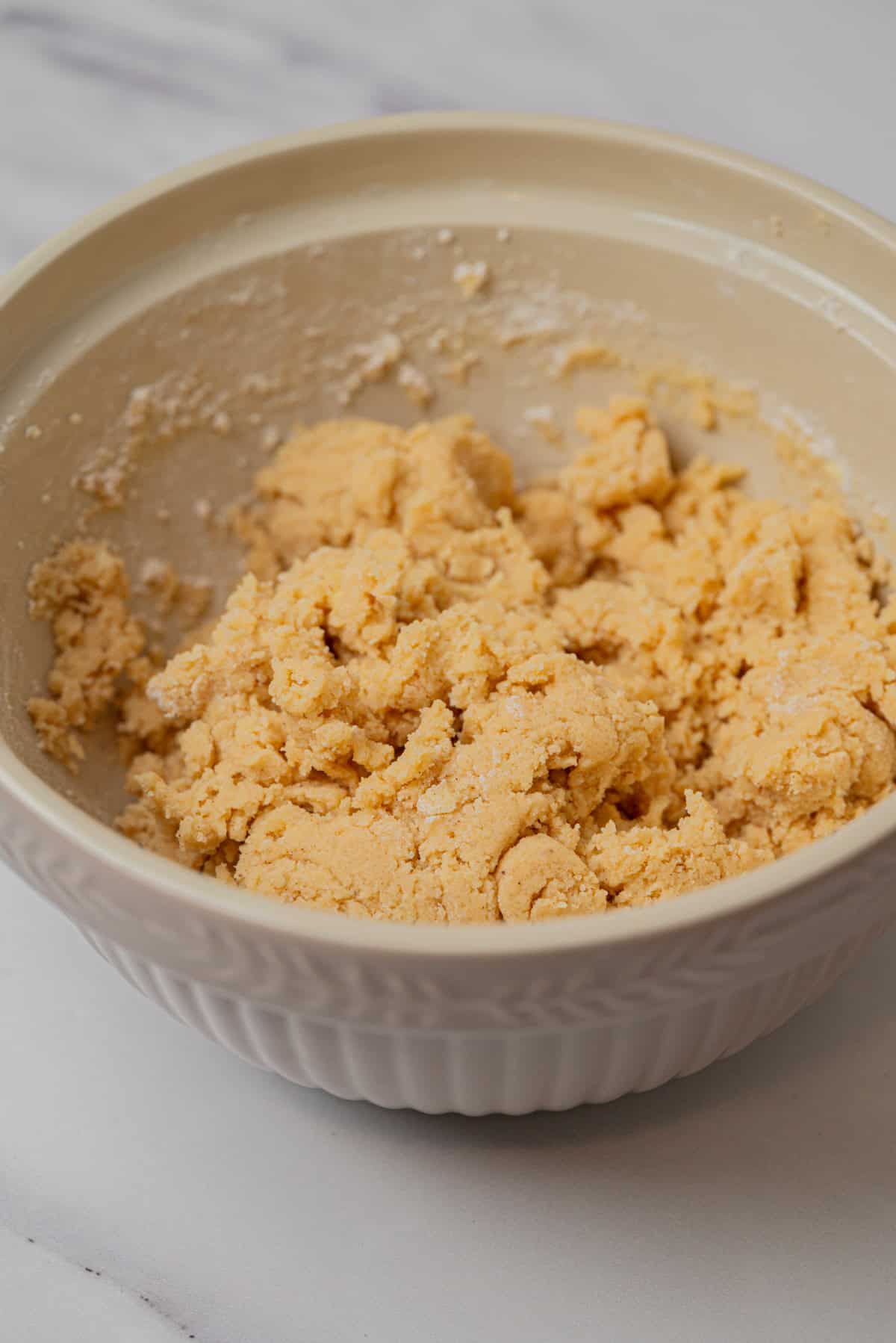 A beige ceramic bowl containing a rough, pale yellow Small-Batch Snickerdoodles cookie dough mixture sits on a light-colored countertop.