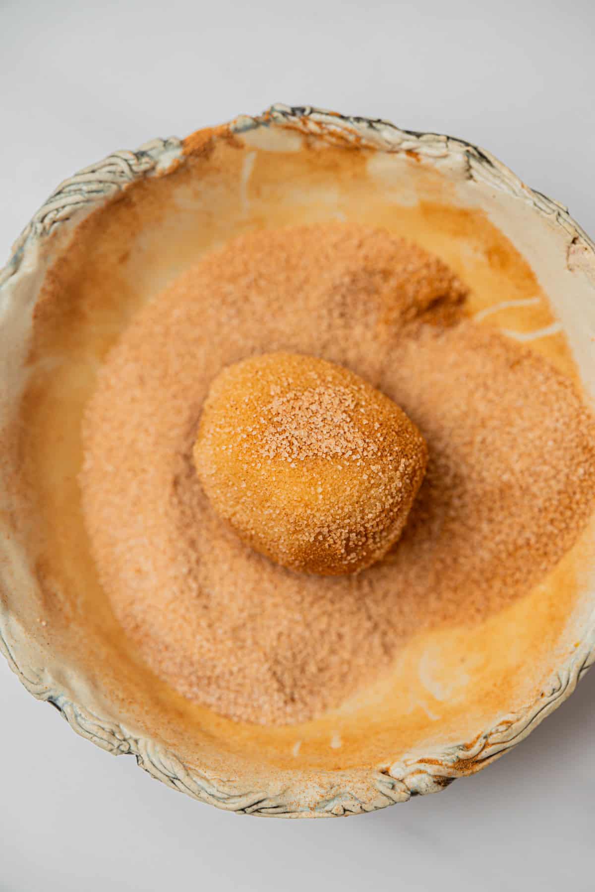 A ball of dough for Small-Batch Snickerdoodles sits in a rustic, shallow bowl filled with cinnamon sugar on a light surface, ready to be transformed into delicious cookies.
