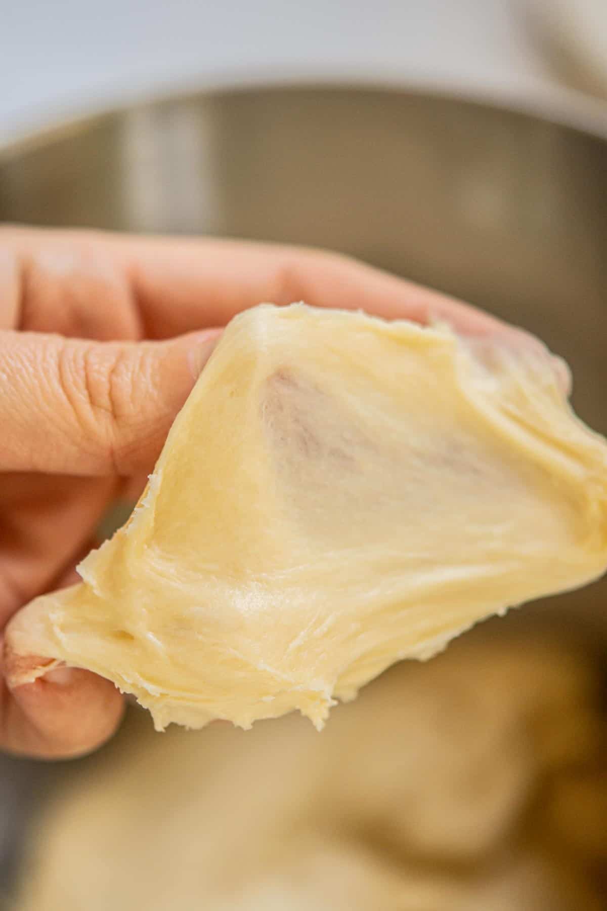 A close-up of a hand gently stretching smooth, elastic dough for small batch sticky buns over a mixing bowl, demonstrating its stretchy texture.