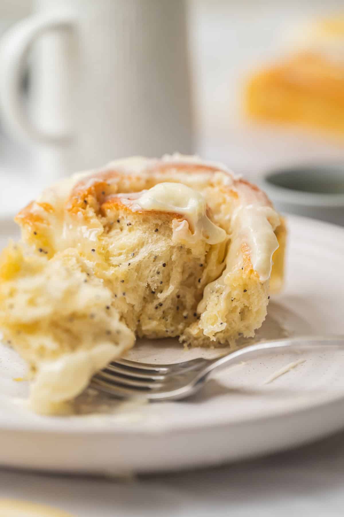 A close-up of a partially eaten frosted cinnamon roll on a white plate, with a fork resting beside it. The cinnamon roll appears soft and fluffy, similar to lemon poppy seed rolls, and is topped with creamy, white icing.