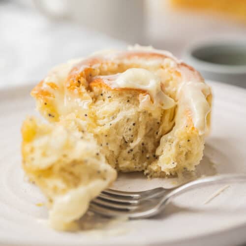 A close-up of a partially eaten cinnamon roll with creamy icing on top, sitting on a white plate with a fork beside it. The soft, fluffy roll shares the plate with lemon poppy seed rolls, showcasing their light and airy texture.