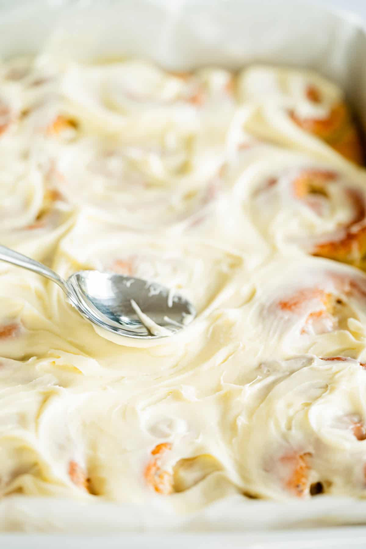 A close-up of a spoon spreading creamy white icing over freshly baked lemon poppy seed rolls in a baking dish.