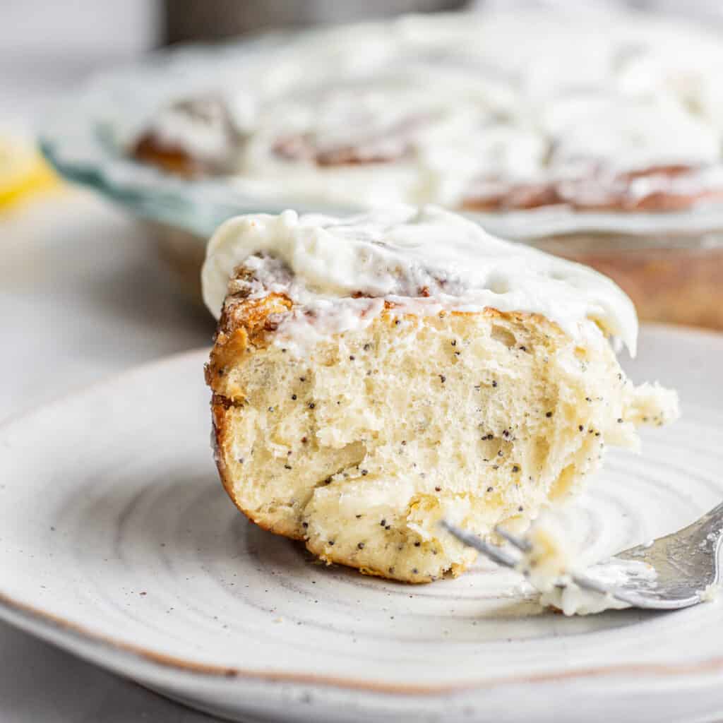 A lemon poppy seed rolls on a plate with a fork and other poppy seed sweet rolls in a tray behind it.