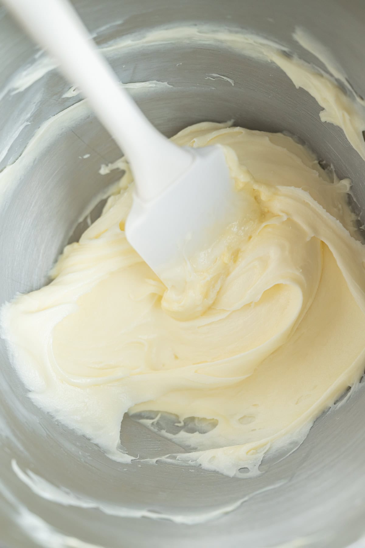 A close-up of a mixing bowl with creamy white frosting being stirred by a white spatula, perfect for topping freshly baked lemon poppy seed rolls.
