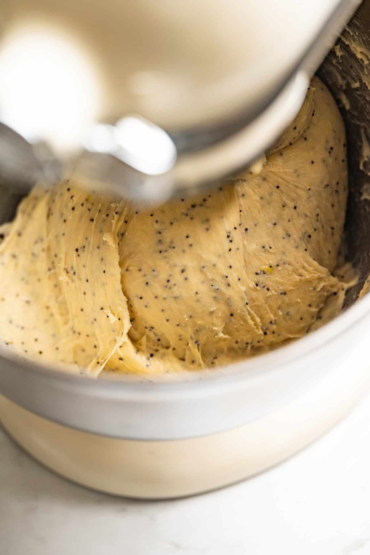 Close-up of a stand mixer bowl filled with creamy lemon poppy seed rolls dough, dotted with poppy seeds and shown mid-mixing.
