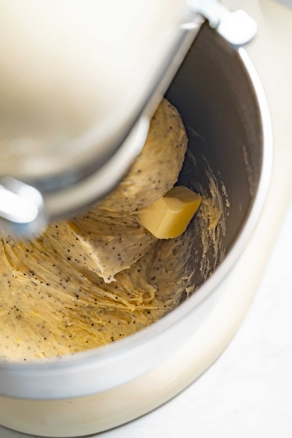 A close-up of a stand mixer bowl with dough for lemon poppy seed rolls, featuring poppy seeds and a chunk of butter being mixed by the machine’s paddle attachment.