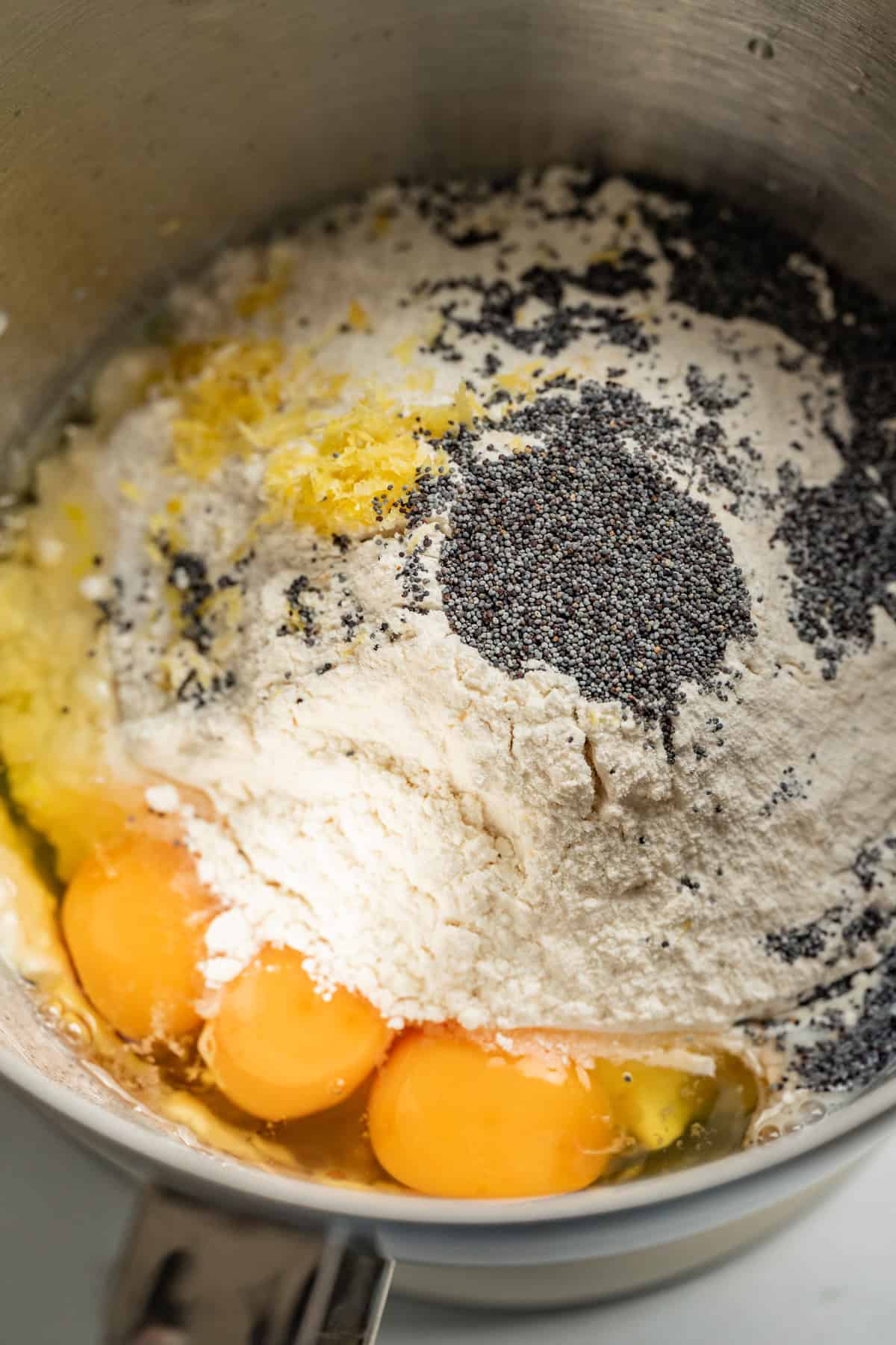 A close-up of a mixing bowl containing flour, eggs, poppy seeds, lemon zest, butter, and sugar, all unmixed and ready for baking delicious lemon poppy seed rolls.