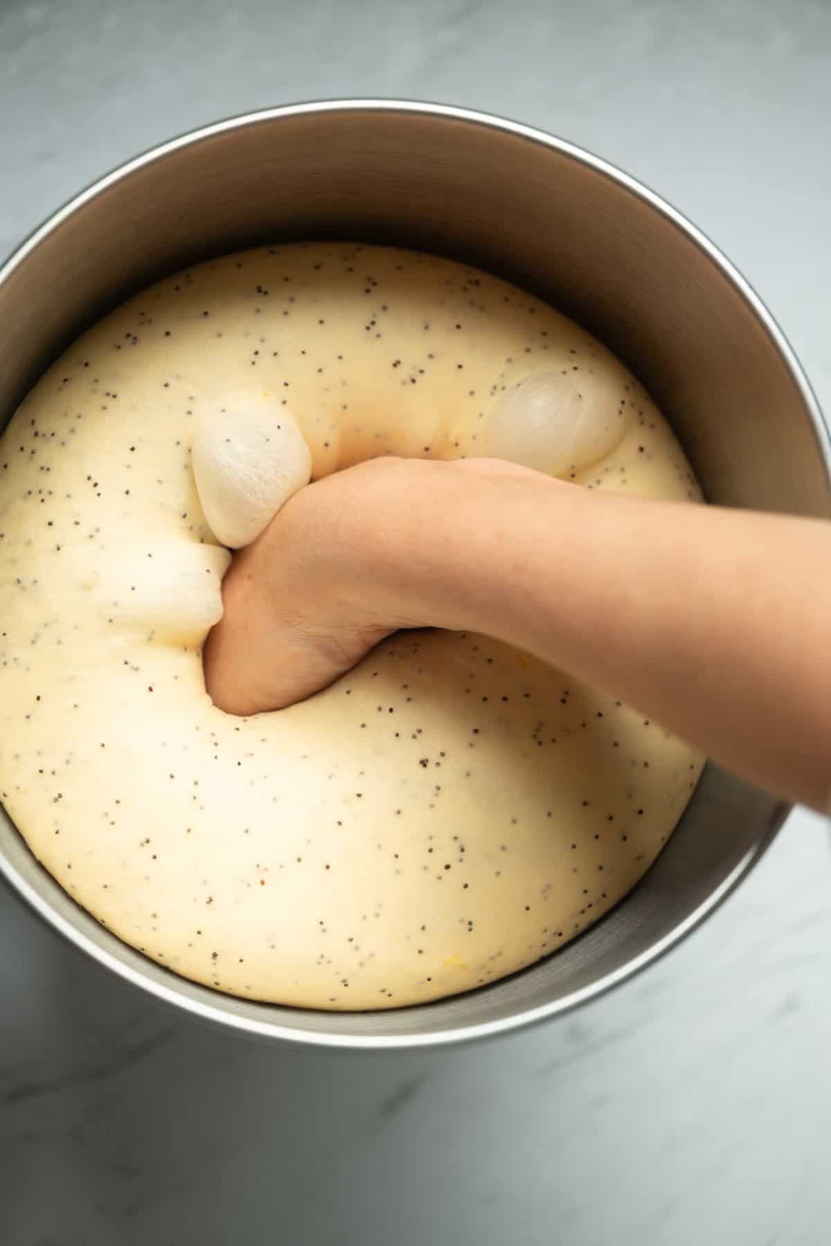 A hand punches down risen lemon poppy seed rolls dough in a large metal mixing bowl on a light gray surface.