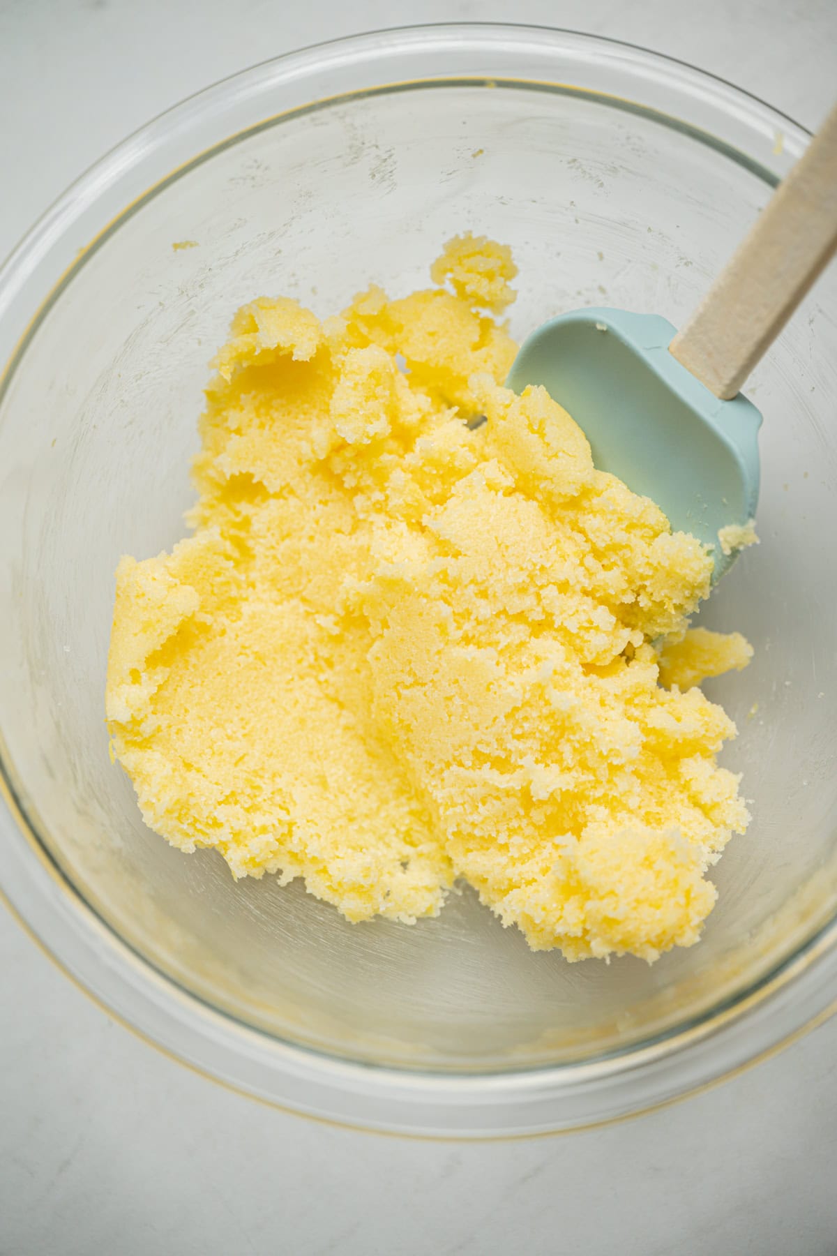 A glass bowl containing a yellow, crumbly mixture for lemon poppy seed rolls, with a light blue silicone spatula resting inside.