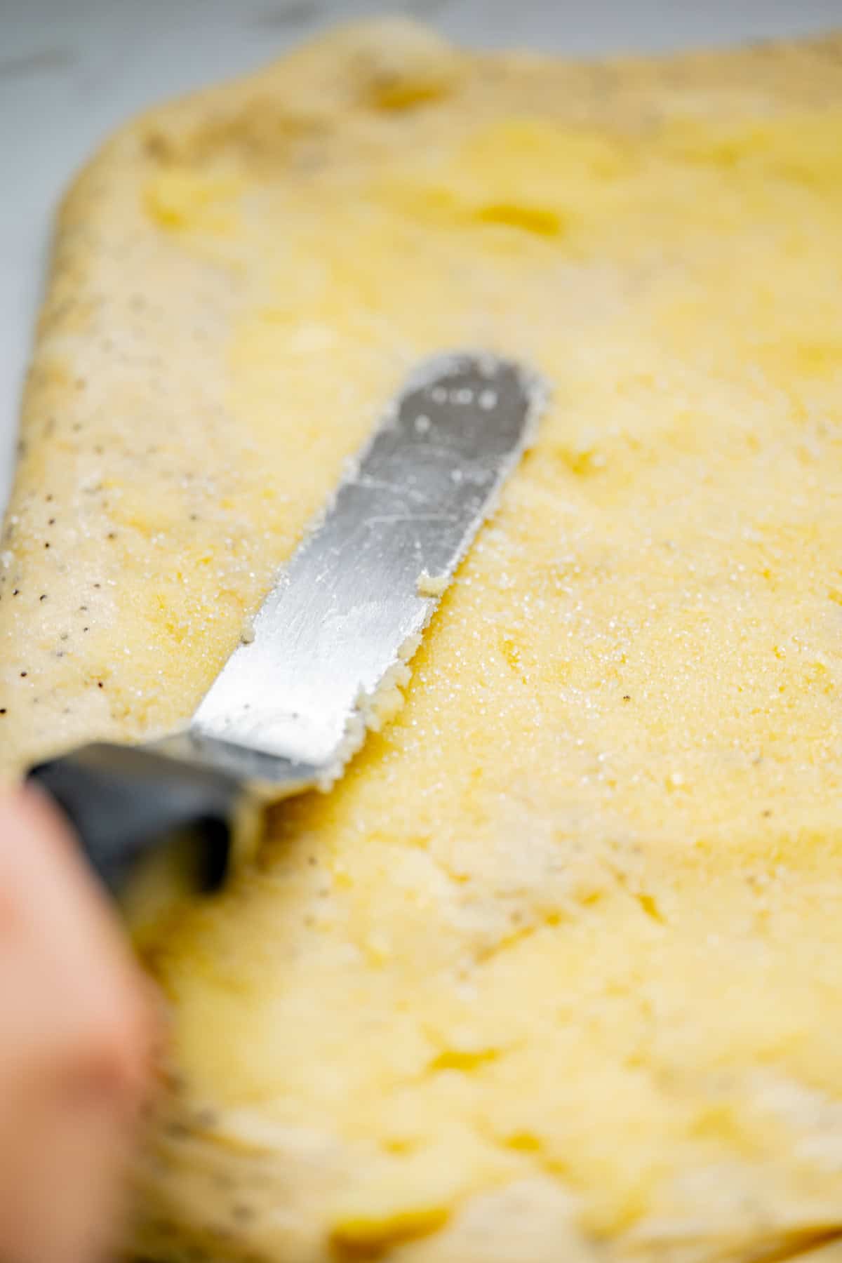 A hand uses an offset spatula to spread a creamy yellow mixture evenly over a sheet of dough, preparing delicious lemon poppy seed rolls for baking.