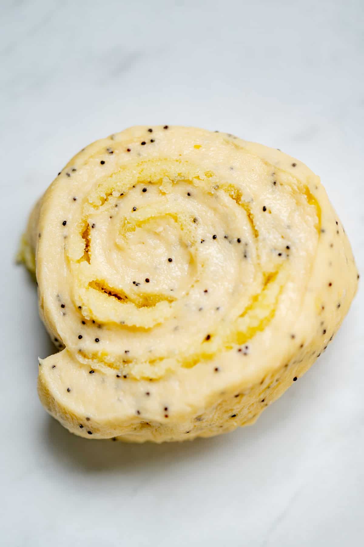 Close-up of an unbaked lemon poppy seed roll with visible seeds in the dough, resting on a light-colored surface. The spiral shape and smooth texture are clearly shown.