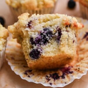 A close-up of a blueberry buttermilk muffin with a bite taken out, revealing its moist, fluffy texture and juicy blueberries inside. The muffin rests on an unwrapped paper liner.