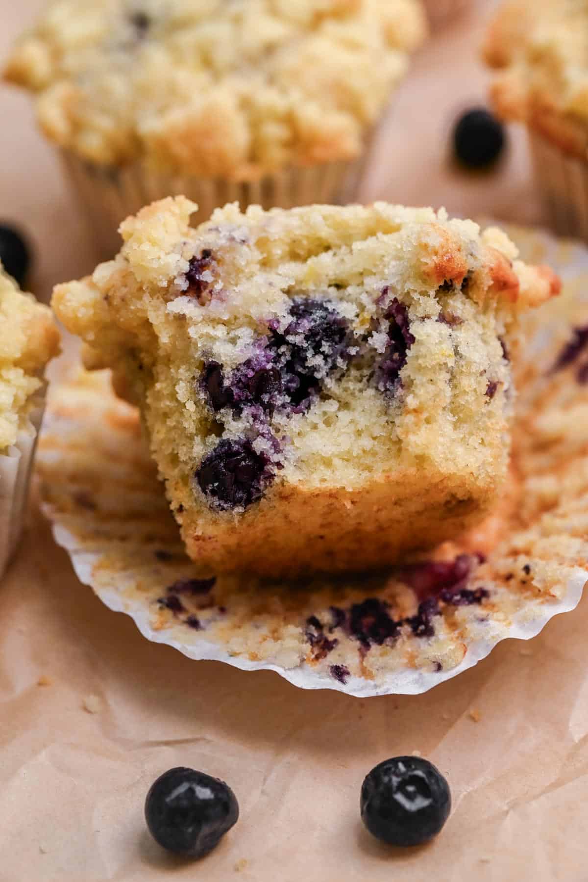 A close-up of a blueberry buttermilk muffin cut in half, showing a moist, fluffy interior dotted with blueberries. The muffin rests on a crinkled paper liner, with fresh blueberries scattered nearby.