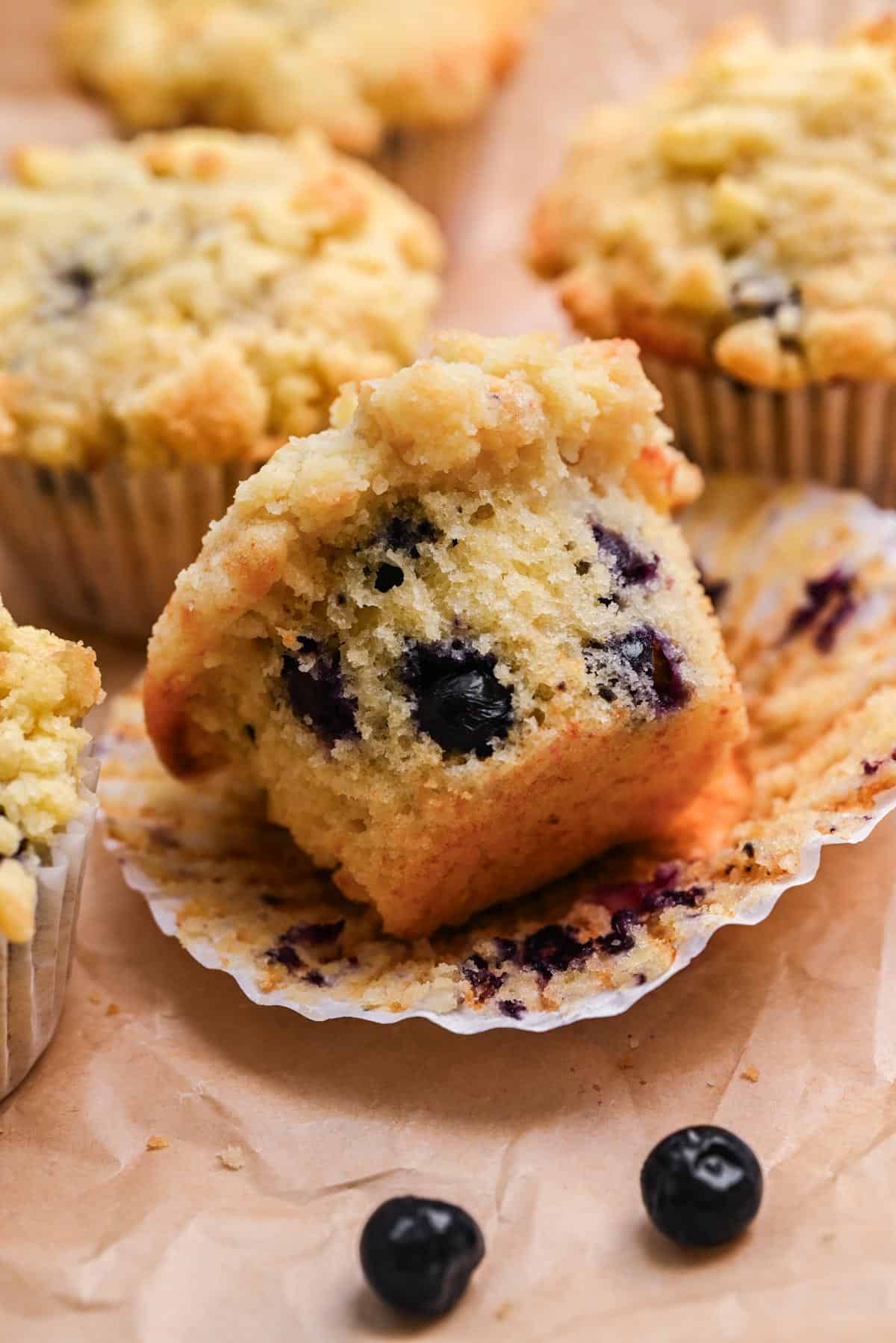 A close-up of a blueberry buttermilk muffin cut in half, revealing juicy blueberries inside. The muffin rests on a crinkled paper liner with a few loose blueberries nearby and more blueberry buttermilk muffins in the background.