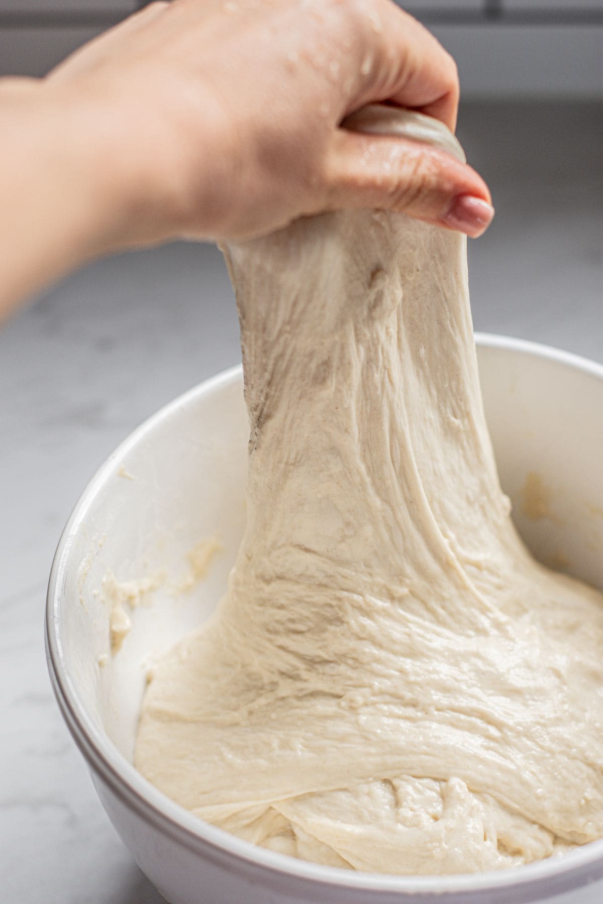 A hand stretches sticky pesto focaccia dough from a white mixing bowl on a light kitchen countertop.