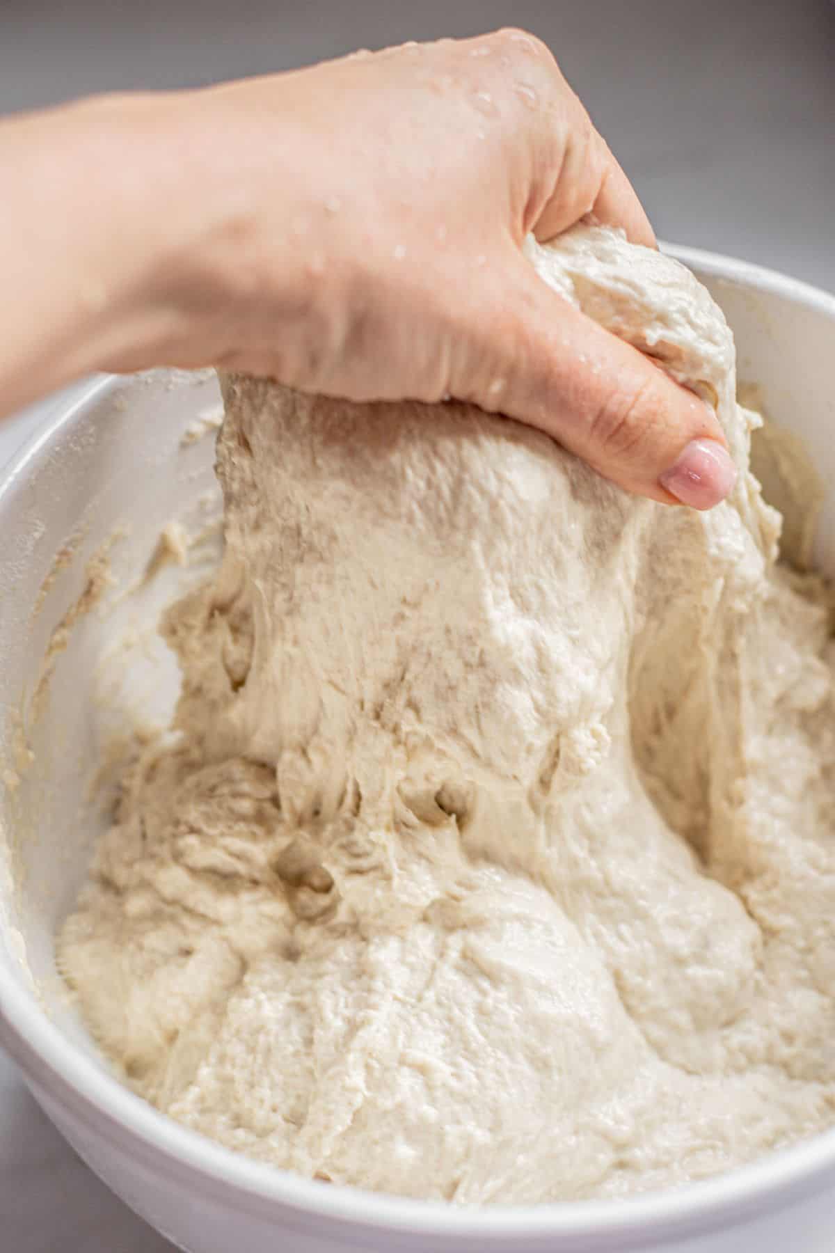 A hand is stretching and kneading sticky pesto focaccia dough in a white mixing bowl. The dough appears soft and elastic, with strands pulling upward from the bowl.