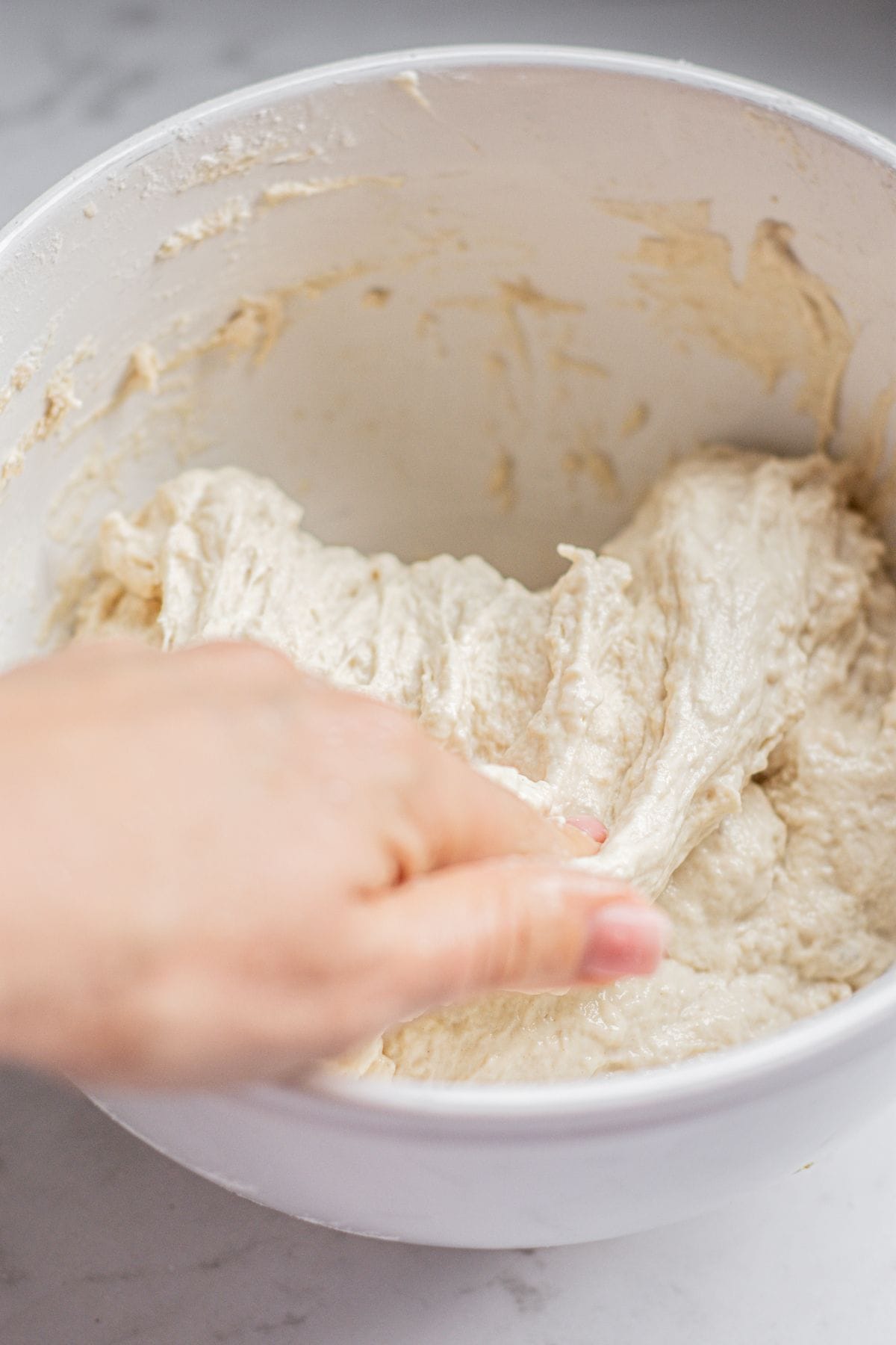 A hand stretches sticky pesto focaccia dough inside a white mixing bowl on a light-colored countertop.