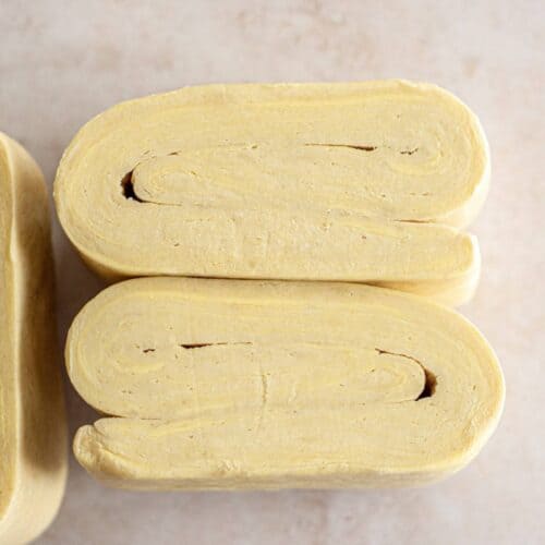 Close-up of folded dough layers on a light background. The rough puff pastry is neatly layered and slightly curved at the edges, showcasing a light beige color with a smooth texture.