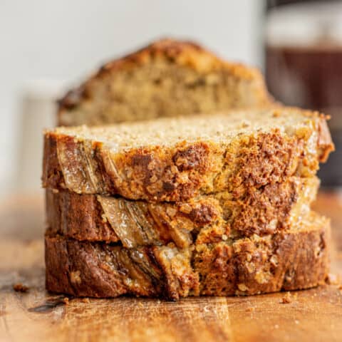 Three thick slices of brown butter banana bread stacked on a wooden surface, showing a moist, dense texture with a golden-brown crust and visible banana pieces.