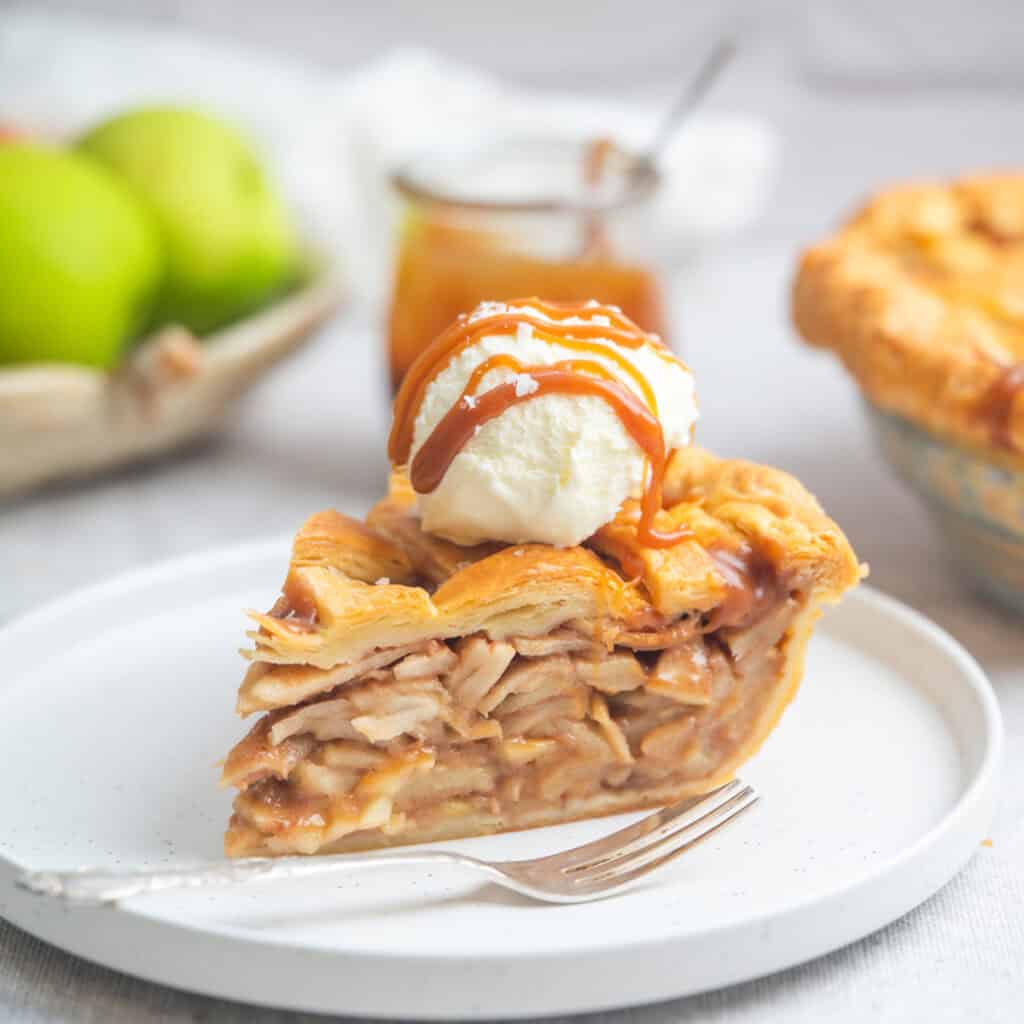 A slice of caramel apple pie topped with vanilla ice cream and caramel sauce sits on a white plate with a fork. In the background, green apples and a jar of caramel sauce add to the delicious scene.