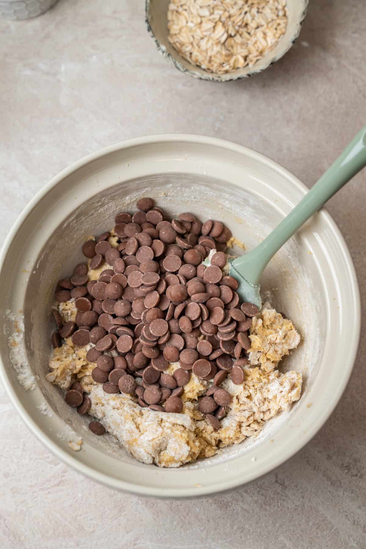 A mixing bowl containing oatmeal chocolate chip cookie dough topped with chocolate chips and a silicone spatula, with a small bowl of oats in the background.