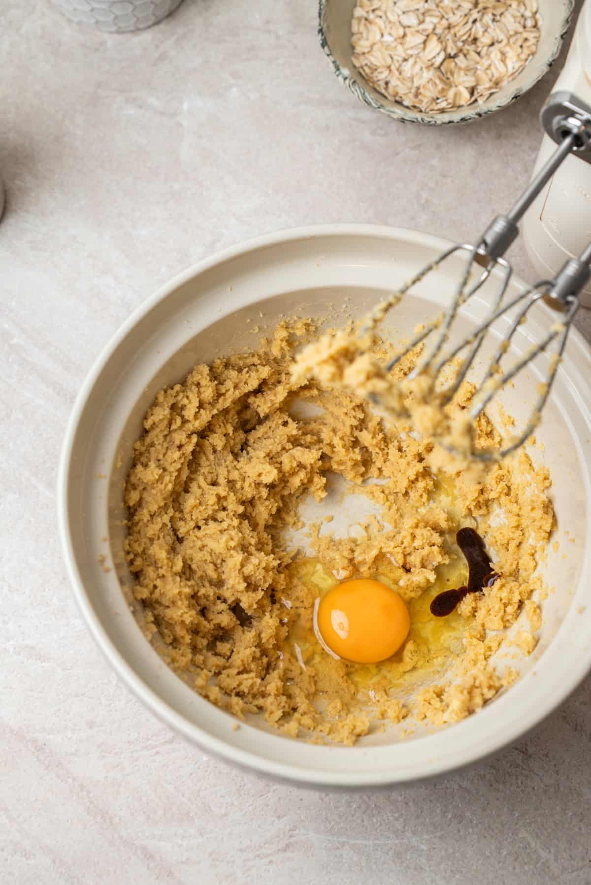 A mixing bowl containing creamed butter and sugar, a cracked egg, and vanilla extract for oatmeal chocolate chip bars, with electric beaters nearby.